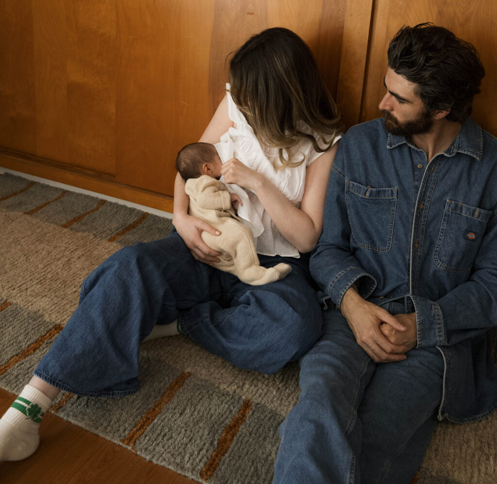 in-home newborn session Paul and Nicole sitting on their mid-century modern couch, Paul holding newborn, editorial-style pose in a warm, wood-paneled room.