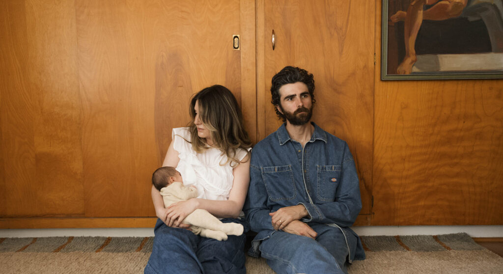 in-home newborn session with Paul and Nicole sitting on their mid-century modern couch, Paul holding newborn, editorial-style pose in a warm, wood-paneled room.