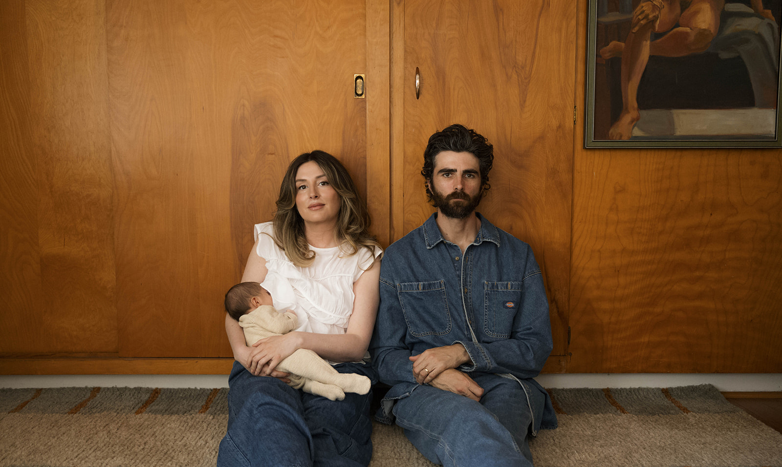 Paul and Nicole sitting on their mid-century modern couch, Paul holding newborn, editorial-style pose in a warm, wood-paneled room.