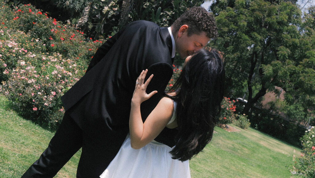 Engagement photo of a couple kissing in a rose garden, with the woman in a short white dress and the man in a black suit, romantic outdoor setting under a clear sky.