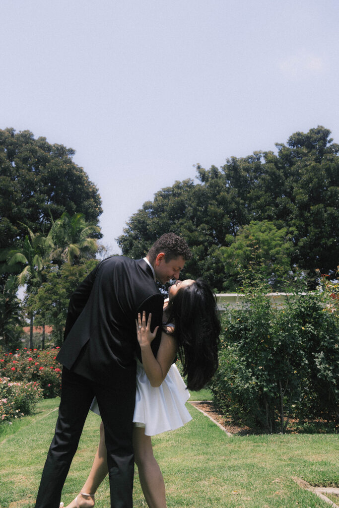 Engagement photo of a couple kissing in a rose garden, with the woman in a short white dress and the man in a black suit, romantic outdoor setting under a clear sky.