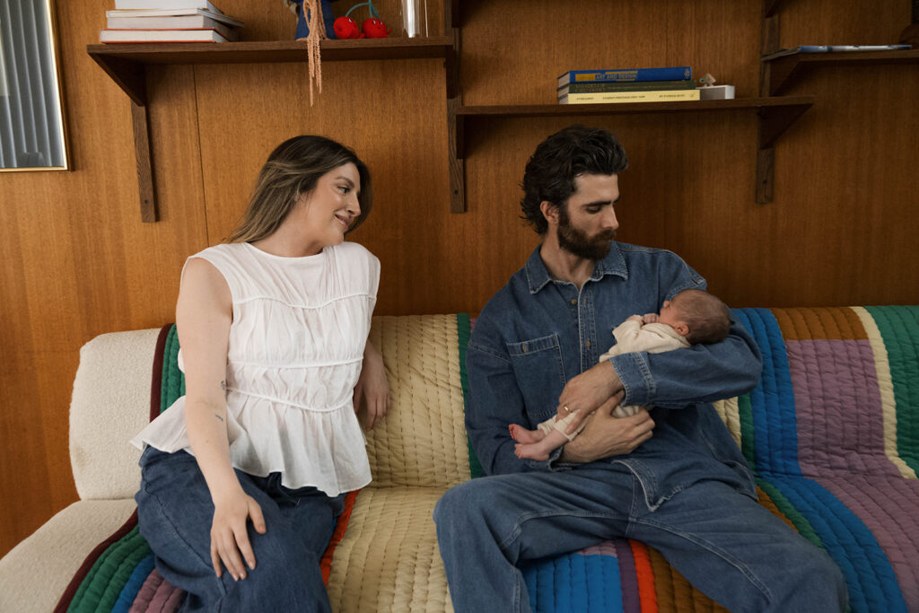 in-home newborn session with Paul and Nicole sitting on their mid-century modern couch, Paul holding newborn, editorial-style pose in a warm, wood-paneled room.