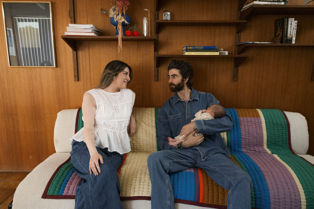 in-home newborn session with Paul and Nicole sitting on their mid-century modern couch, Paul holding newborn, editorial-style pose in a warm, wood-paneled room.