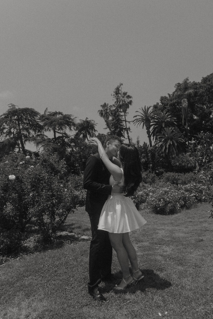 Engagement photo of a couple kissing in a rose garden, with the woman in a short white dress and the man in a black suit, romantic outdoor setting under a clear sky.