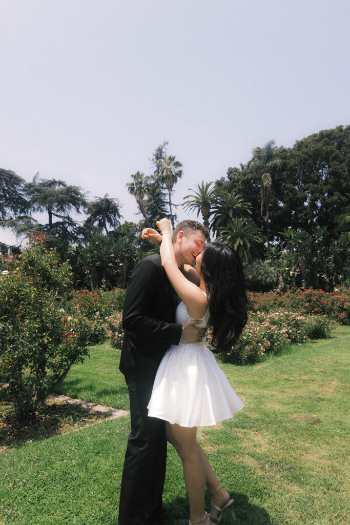Engagement photo of a couple kissing in a rose garden, with the woman in a short white dress and the man in a black suit, romantic outdoor setting under a clear sky.