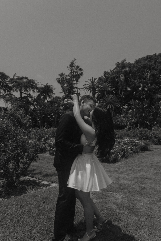 Engagement photo of a couple kissing in a rose garden, with the woman in a short white dress and the man in a black suit, romantic outdoor setting under a clear sky.