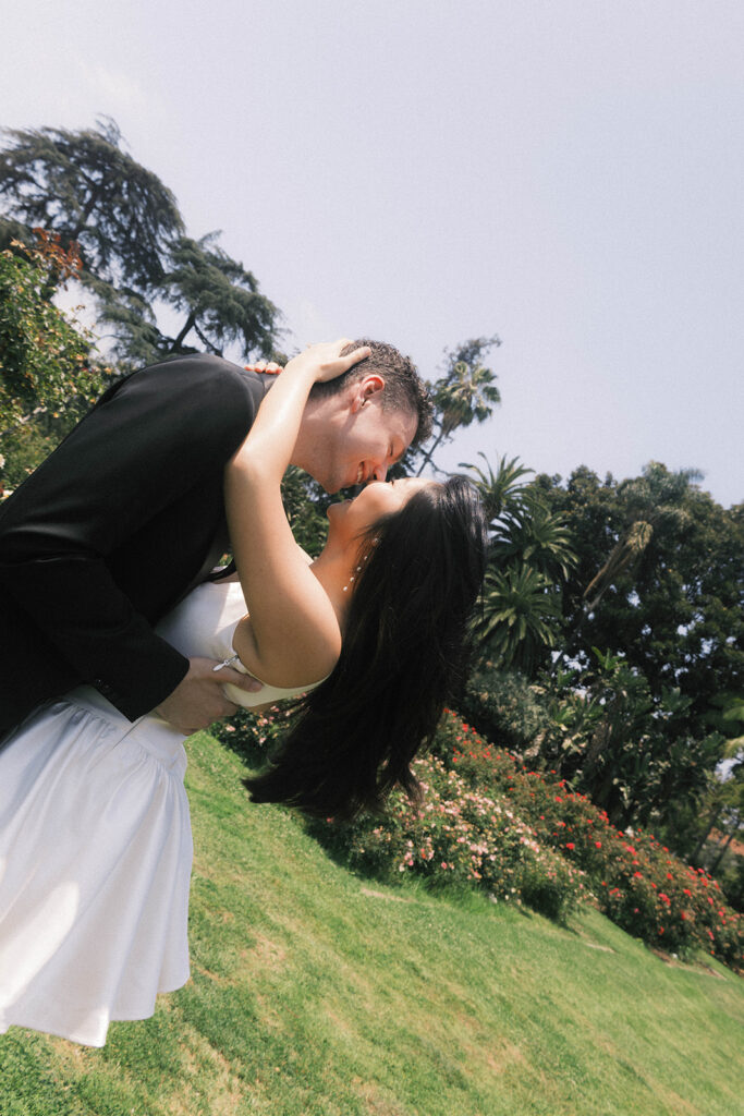 Engagement photo of a couple kissing in a rose garden, with the woman in a short white dress and the man in a black suit, romantic outdoor setting under a clear sky.
