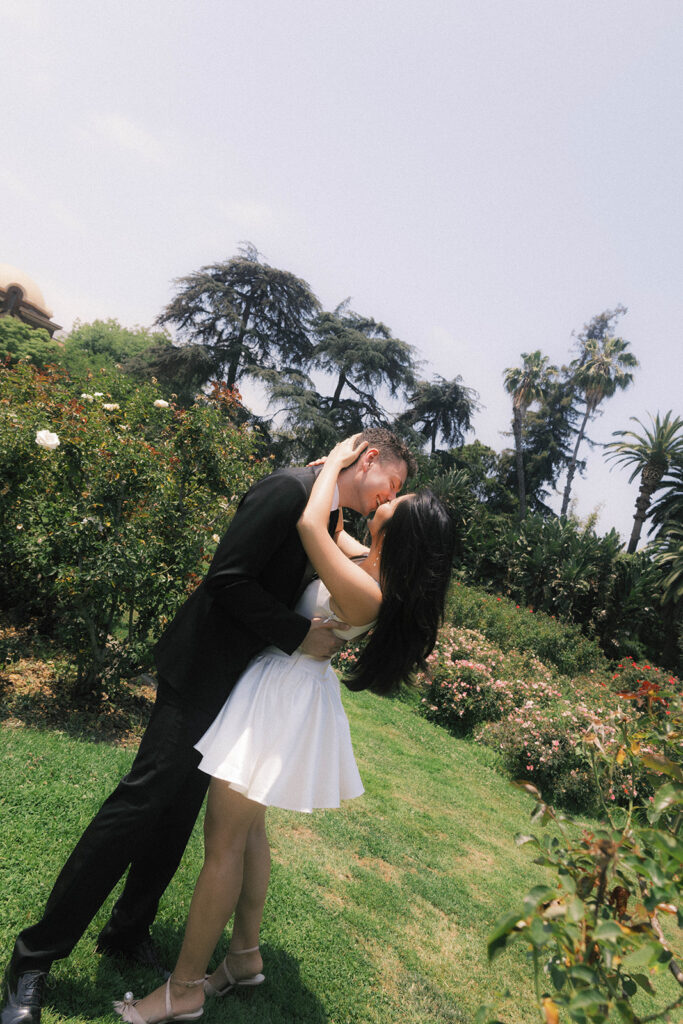 Engagement photo of a couple kissing in a rose garden, with the woman in a short white dress and the man in a black suit, romantic outdoor setting under a clear sky.