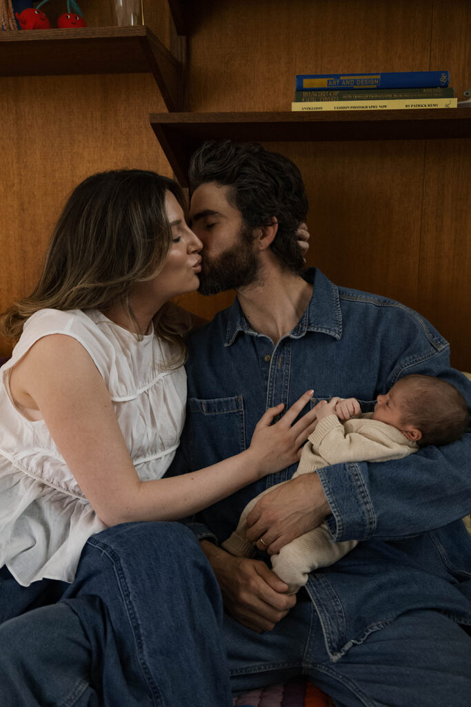in-home newborn session with Paul and Nicole sitting on their mid-century modern couch, Paul holding newborn, editorial-style pose in a warm, wood-paneled room.