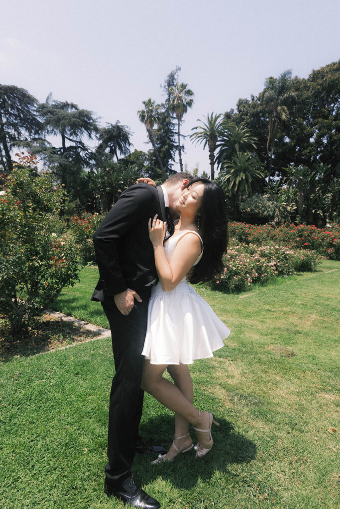 Engagement photo of a couple kissing in a rose garden, with the woman in a short white dress and the man in a black suit, romantic outdoor setting under a clear sky.