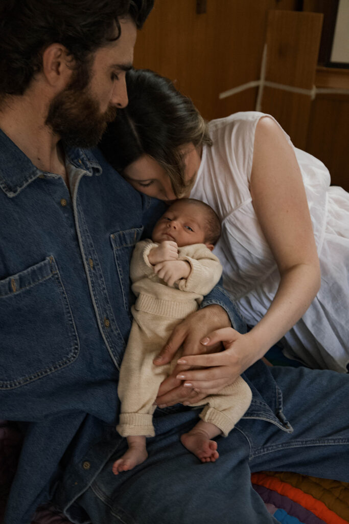 in-home newborn session with Paul and Nicole sitting on their mid-century modern couch, Paul holding newborn, editorial-style pose in a warm, wood-paneled room.