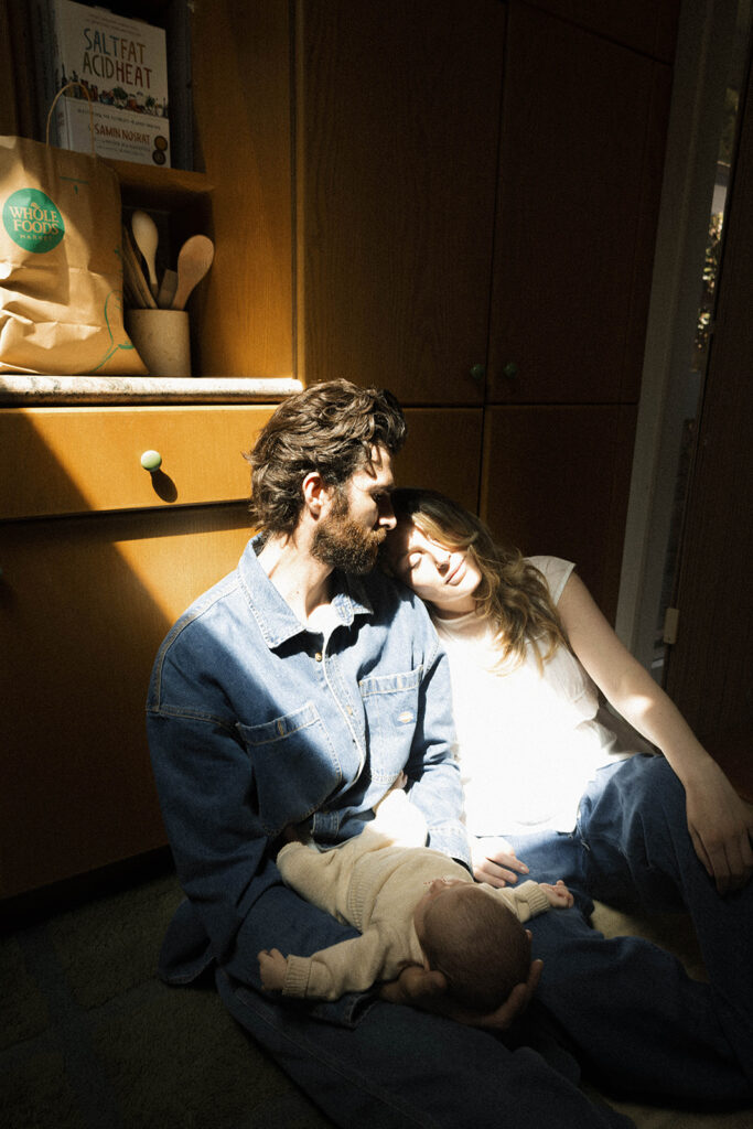 Paul and Nicole sitting on the kitchen floor in soft sunlight, newborn lying across Paul’s lap, shadows and light playing across their faces.