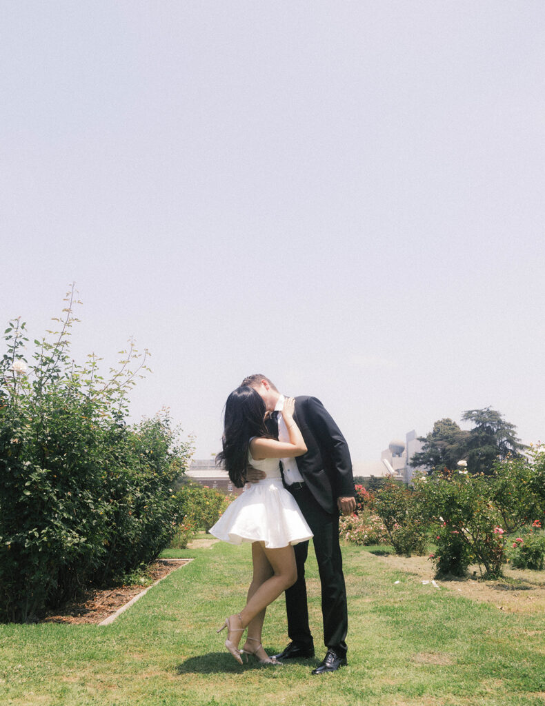 Engagement photo of a couple kissing in a rose garden, with the woman in a short white dress and the man in a black suit, romantic outdoor setting under a clear sky.