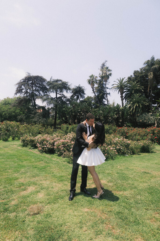 Engagement photo of a couple kissing in a rose garden, with the woman in a short white dress and the man in a black suit, romantic outdoor setting under a clear sky.