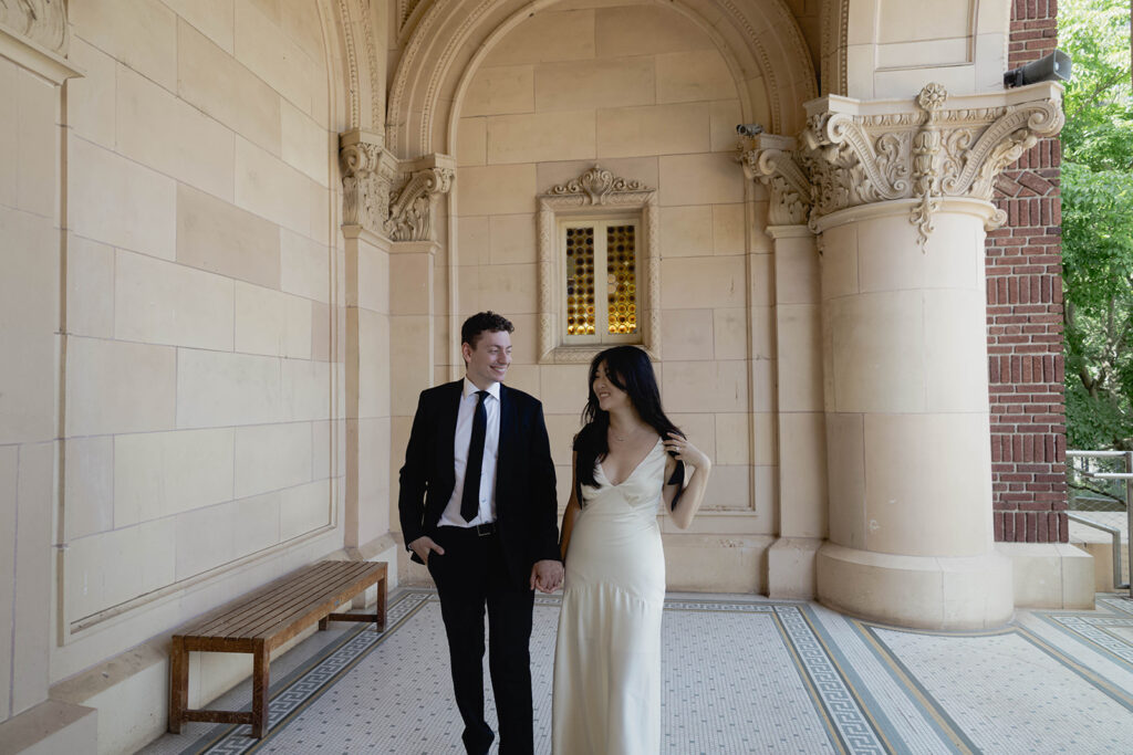 Engagement photo of a couple at natural history museum, with the woman in a long ivory dress and the man in a black suit, romantic outdoor setting under a clear sky.