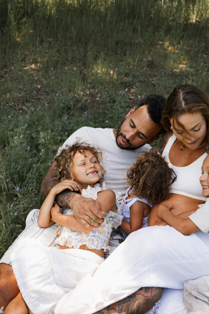Family of five sitting in a grassy area under a tree, children touching and hugging mother's pregnant belly during a candid maternity session in nature

