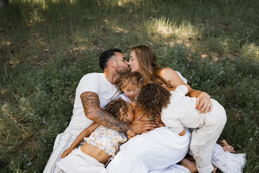 Family of five sitting in a grassy area under a tree, children touching and hugging mother's pregnant belly during a candid maternity session in nature

