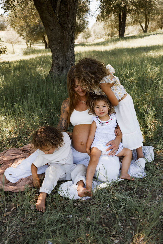 Family of five sitting in a grassy area under a tree, children touching and hugging mother's pregnant belly during a candid maternity session in nature

