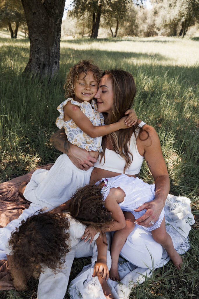Family of five sitting in a grassy area under a tree, children touching and hugging mother's pregnant belly during a candid maternity session in nature

