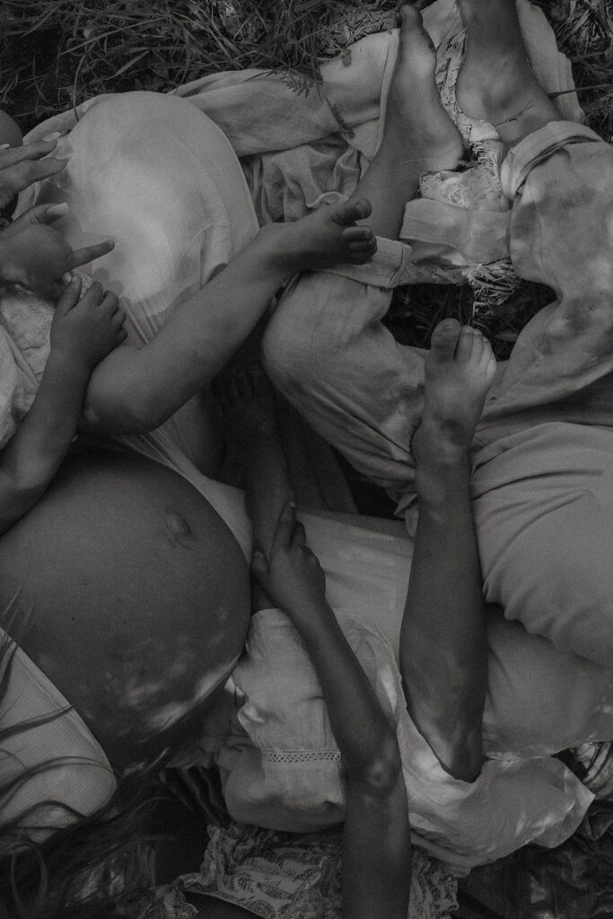 Family of five sitting in a grassy area under a tree, children touching and hugging mother's pregnant belly during a candid maternity session in nature


