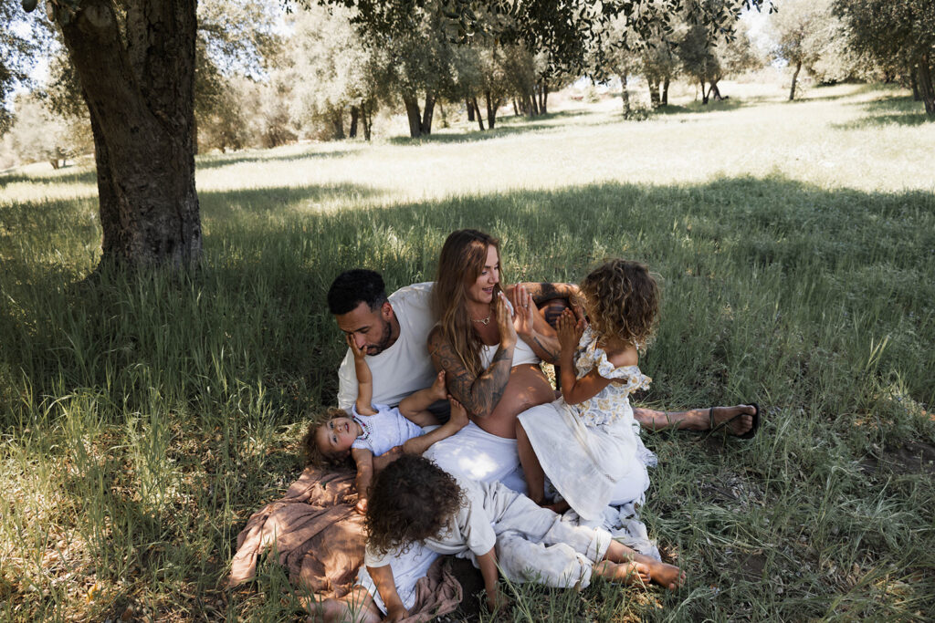 Family of five sitting in a grassy area under a tree, children touching and hugging mother's pregnant belly during a candid maternity session in nature

