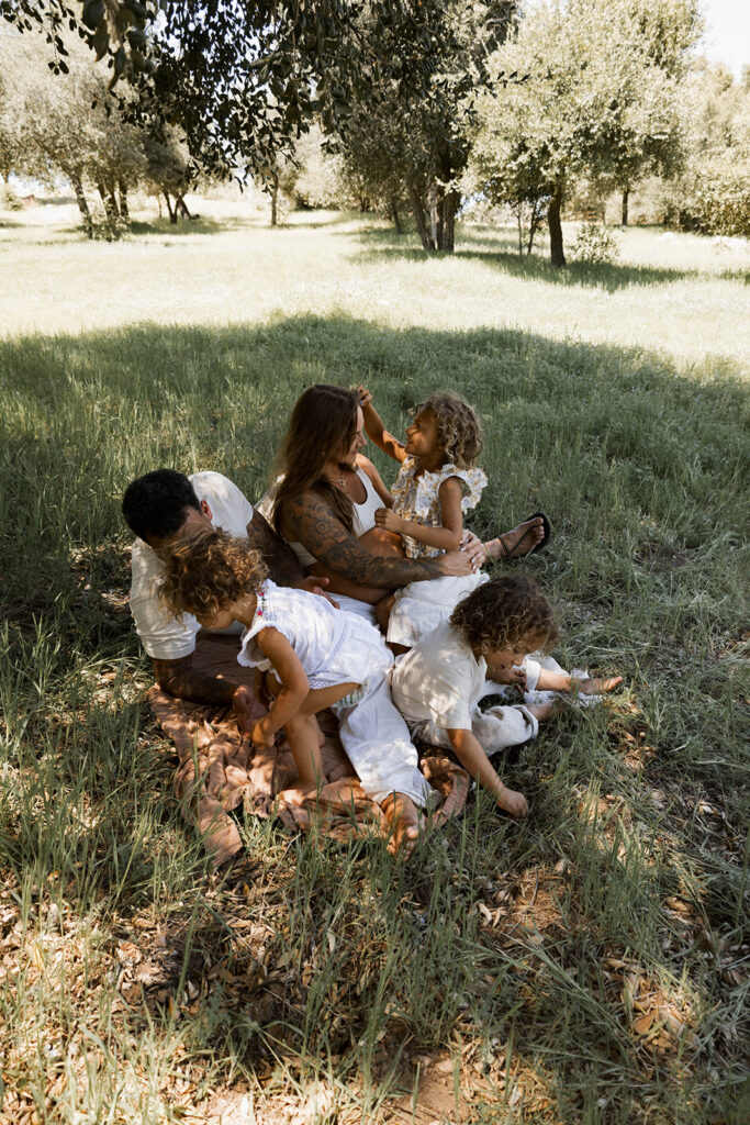 Family of five sitting in a grassy area under a tree, children touching and hugging mother's pregnant belly during a candid maternity session in nature

