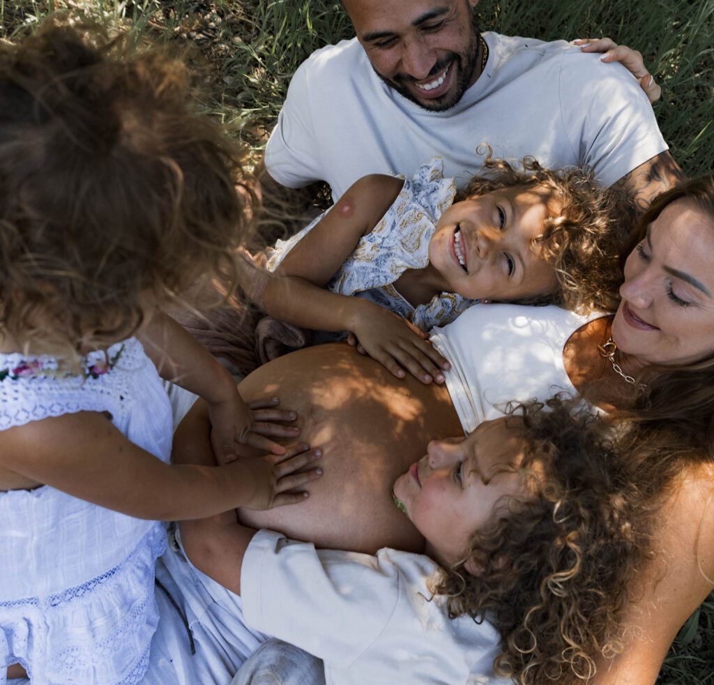 Family of five sitting in a grassy area under a tree, children touching and hugging mother's pregnant belly during a candid maternity session in nature

