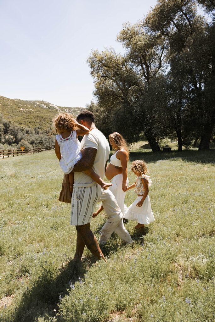 Family walking through a grassy field in a San Diego park during a maternity session, with pregnant mother holding hands with daughter and father carrying another child