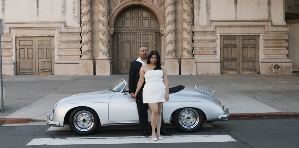 Sabrina and Azor posing in front of a classic vintage car from SD Vintage Rides at Balboa Park, exuding formal glamour.