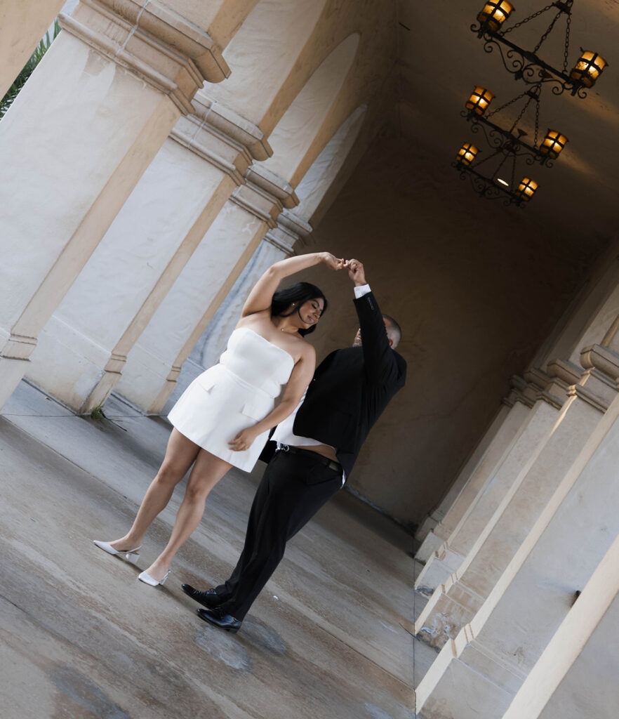 Engaged couple walking hand in hand under the Spanish Colonial arches of Balboa Park, dressed in chic formal attire.