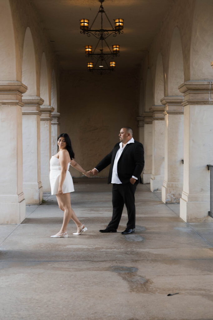 Engaged couple walking hand in hand under the Spanish Colonial arches of Balboa Park, dressed in chic formal attire.