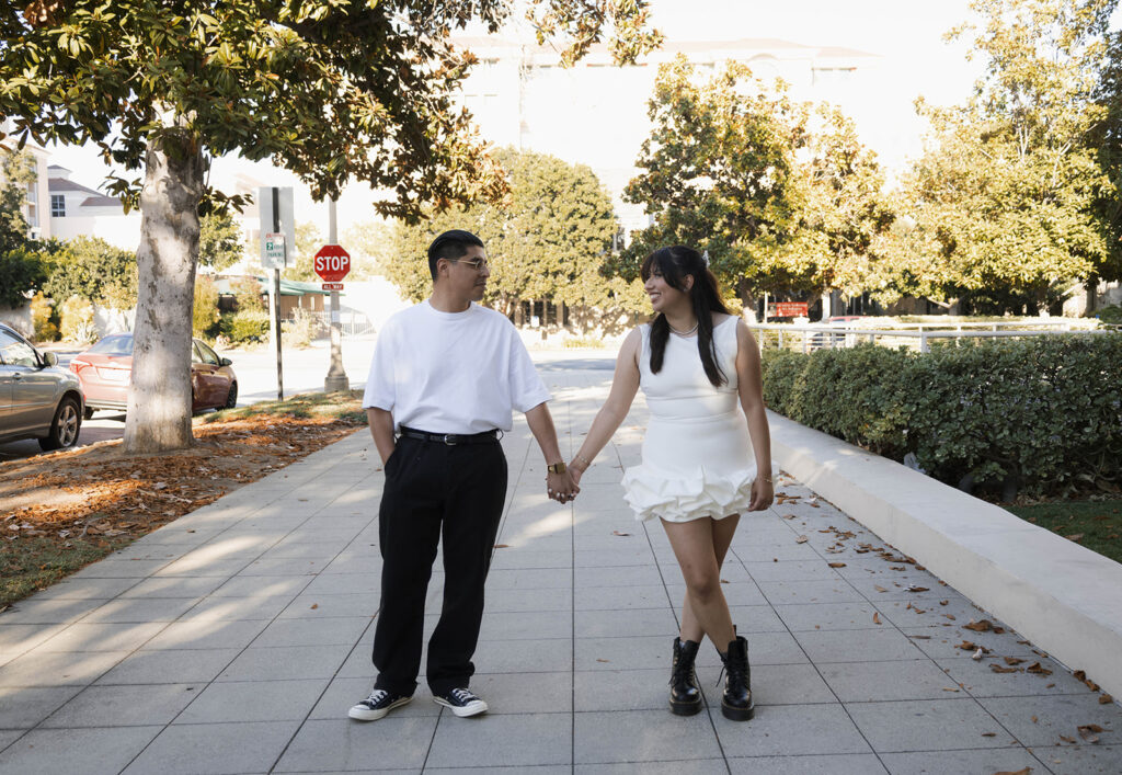 Chris and Andrea share a playful moment walking on a Pasadena sidewalk during their engagement session, with Andrea in a white ruffle dress and black combat boots.