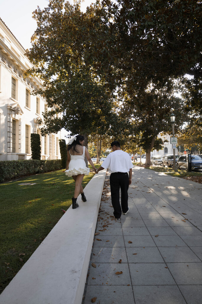 Chris and Andrea share a playful moment walking on a Pasadena sidewalk during their engagement session, with Andrea in a white ruffle dress and black combat boots.