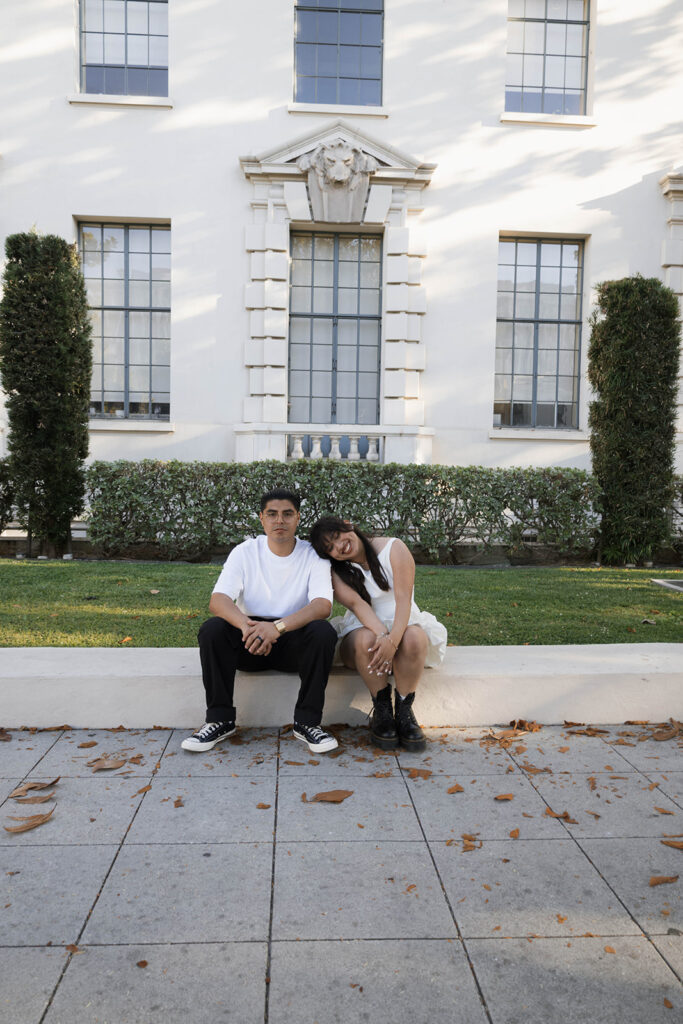 Chris kisses Andrea’s forehead as she rests her head on his shoulder during their Pasadena engagement session, sitting peacefully on a lawn in front of classic architecture.