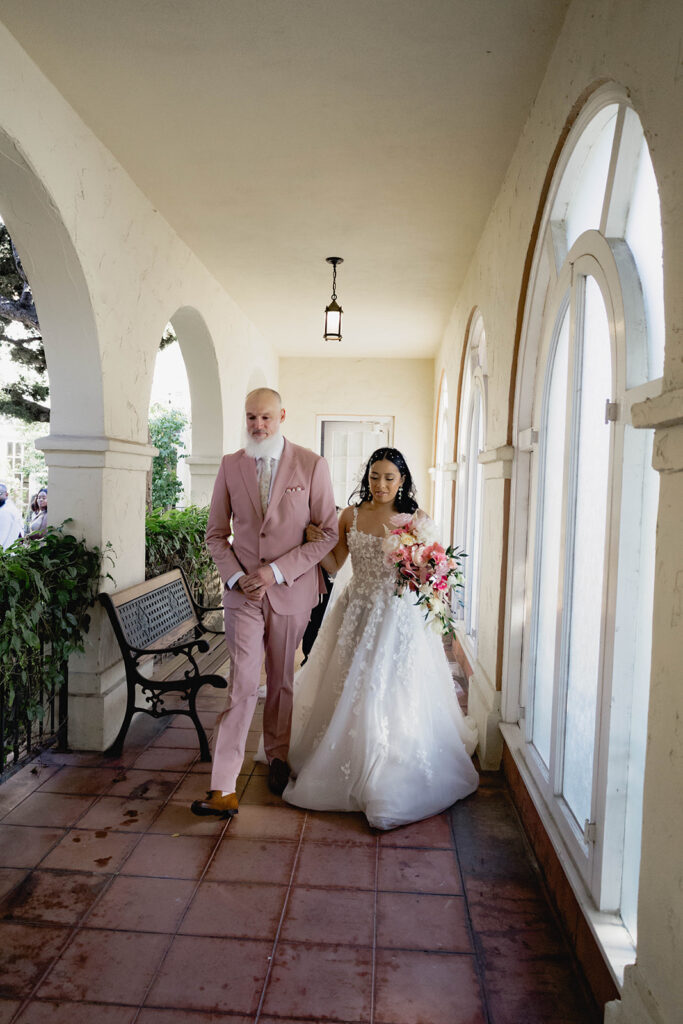 A bride and father walking together to the alter in a courtyard in formal wedding attire.