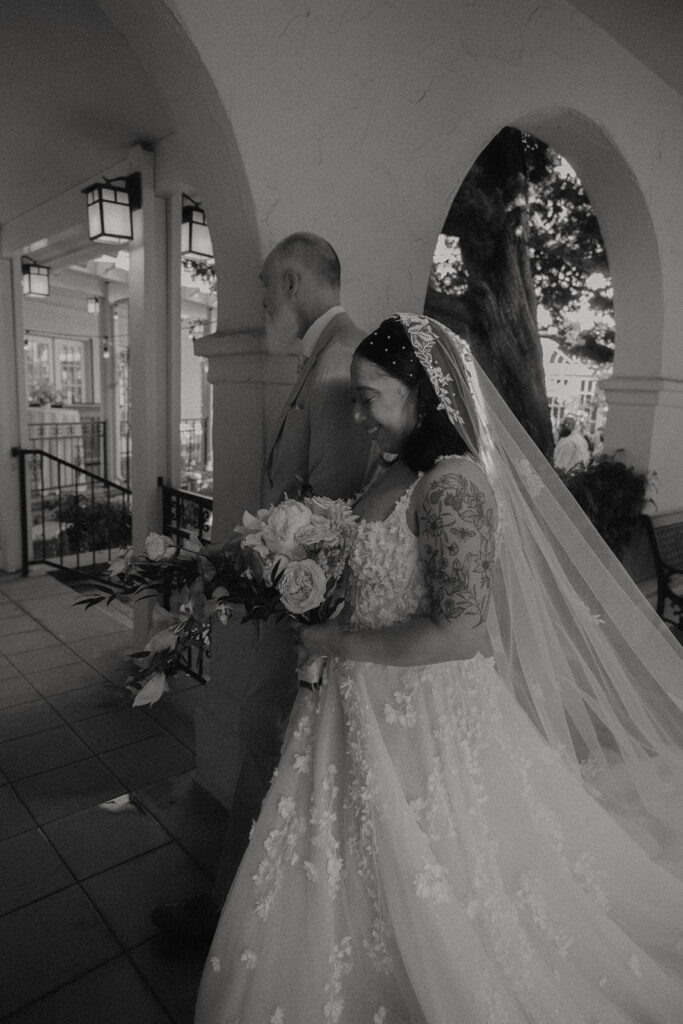 A bride and father walking together to the alter in a courtyard in formal wedding attire.