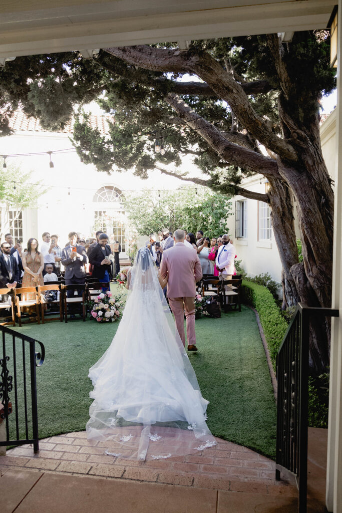 A bride and father walking together to the alter in a courtyard in formal wedding attire.