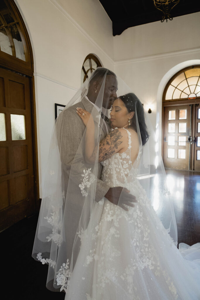 A bride and groom standing together indoors in formal wedding attire kissing.