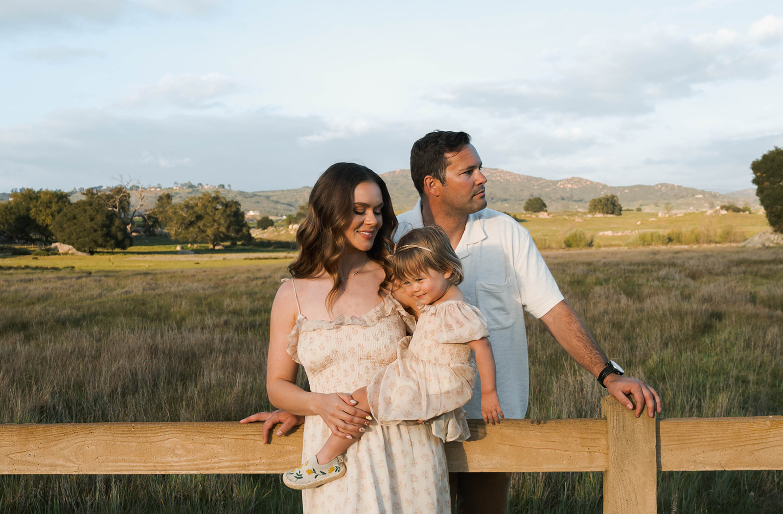 family of 3 taking family photo against wooden fence in a field of grass
